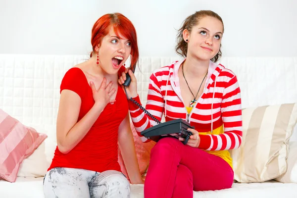 Funny girl sitting on sofa and holding phone at ear her surprised girlfrien