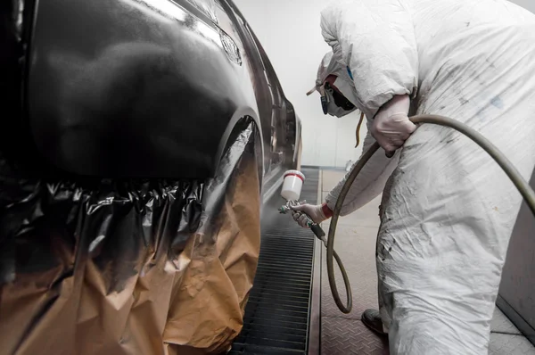 Auto worker spraying black paint on a car in an auto garage with protective gear