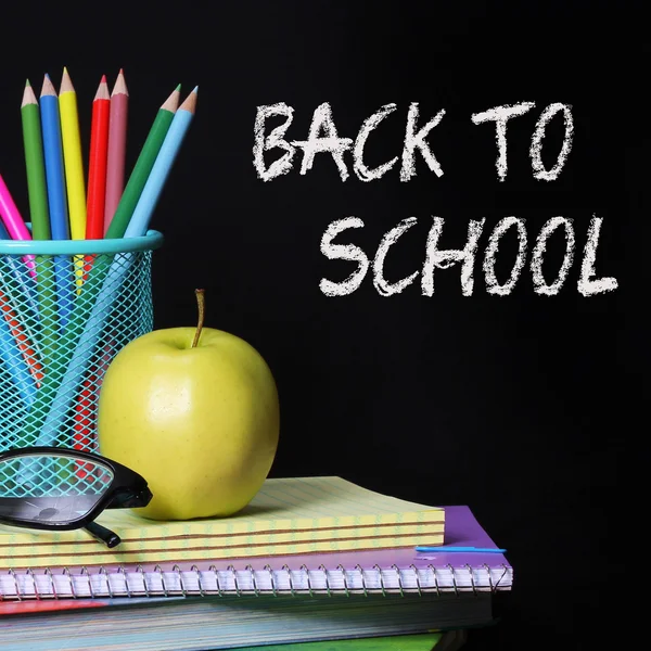 Back to school concept. An apple, colored pencils and glasses on pile of books over black background . The words \'Back to School\' written in chalk on the blackboard
