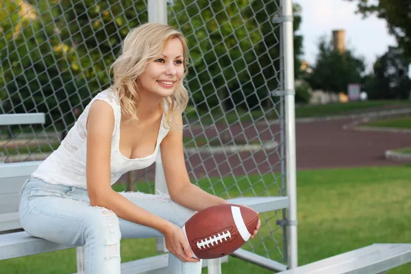 Happy blonde girl with american football. Smiling cheerful beautiful young woman sitting on the bench. Outdoors. Fan of football team is watching the game
