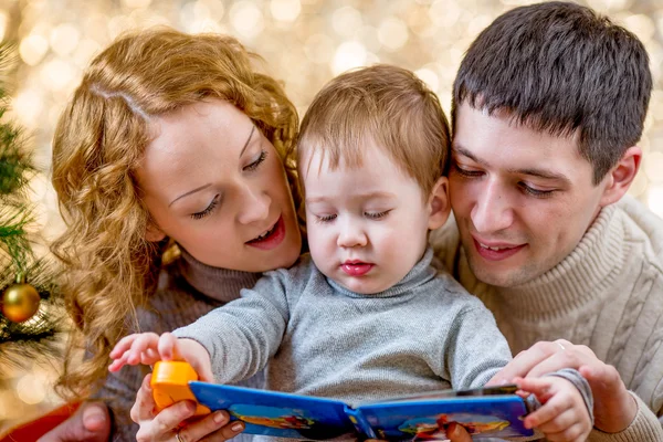 Family at christmas tree. Parents read book to kid.