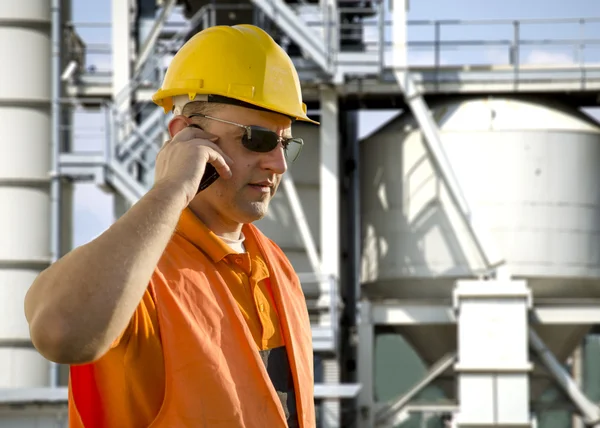 Worker with helmet and sunglasses talking on mobile phone in front of oil plant