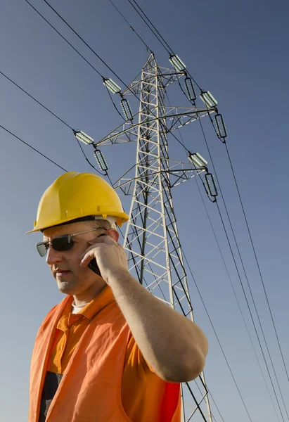 Worker with helmet and sunglasses talking on mobile phone in front of transmission tower