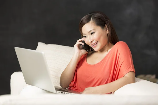 Chinese woman sitting on sofa using laptop and talking on phone.