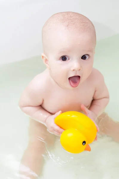 Little smiling baby with yellow duck sitting in white bathtub