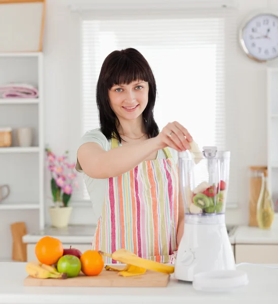 Beautiful brunette woman putting vegetables in a mixer while sta