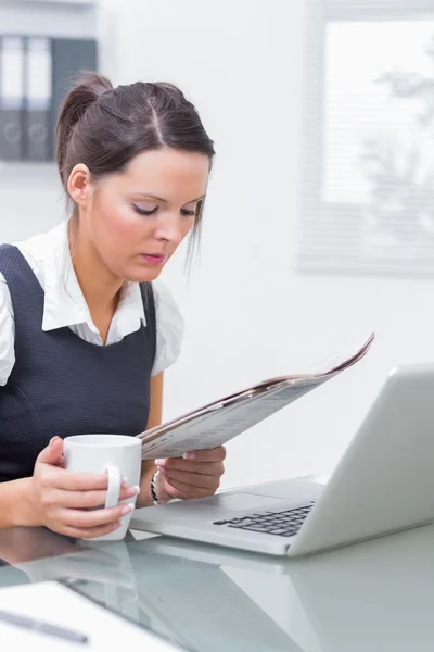 Business woman with coffee cup and laptop reading paper at offic