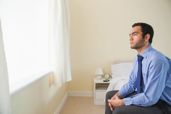 Unsmiling man looking out the window sitting on his bed