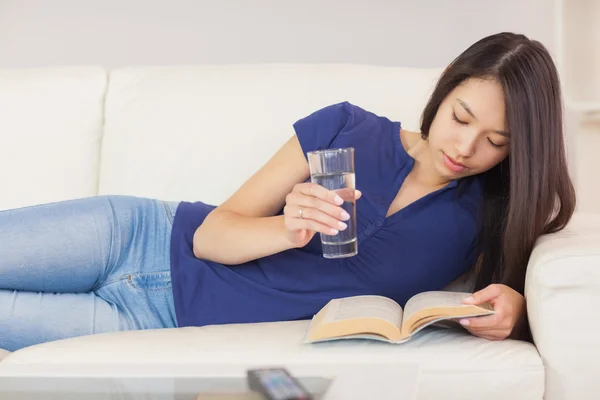 Happy asian girl lying on the sofa reading a novel and drinking water