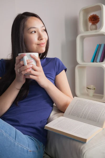 Thinking asian woman sitting on the couch holding mug of coffee