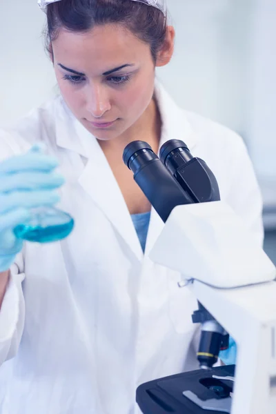 Female researcher examining blue liquid in flask by microscope i