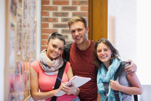 Three smiling students standing next to notice board