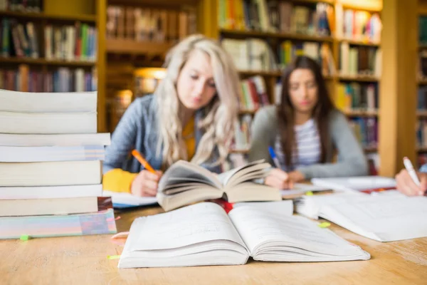 Students writing notes with stack of books at library desk
