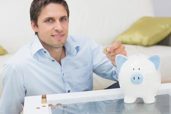 Casual man putting some coins into a piggy bank in living room