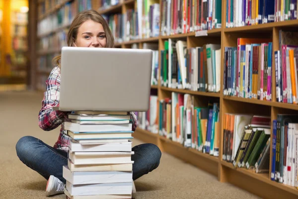 Happy student sitting on library floor using laptop on pile of books
