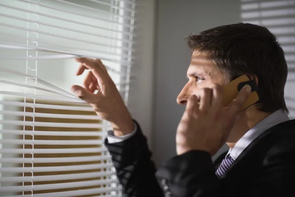 Businessman peeking through blinds while on call in office