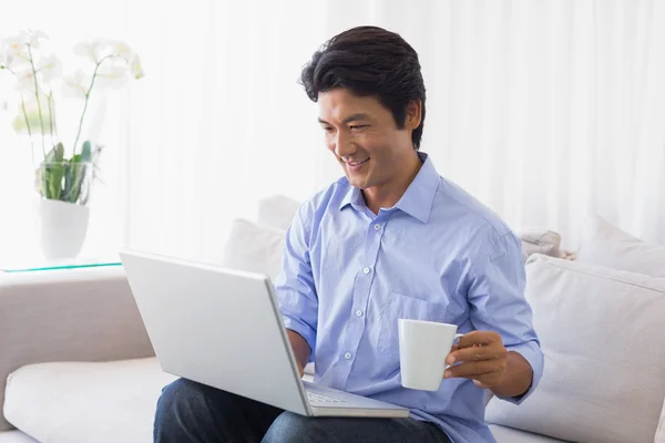 Happy man sitting on couch using laptop having coffee