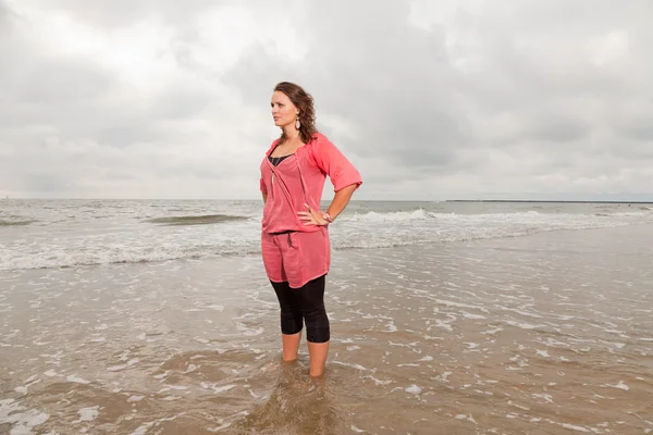 Pretty young woman enjoying outdoor nature near the beach. Standing in the water. Brown hair. Wearing pink shirt. Cloudy sky.