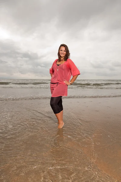 Happy young woman enjoying outdoor nature near the beach. Standing in the water. Brown hair. Wearing pink shirt. Cloudy sky.