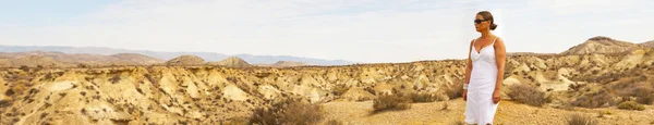 Panoramic photo of tourist brunette woman wearing sunglasses wearing white dress viewing stunning desert landscape with mountains and blue sky. Desierto de Tabernas, Almeria. Andalusia. Spain.