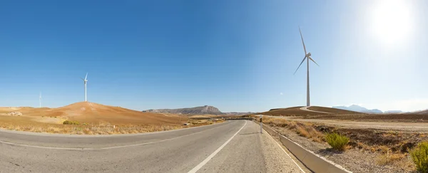 Beautiful panoramic photo of road through agrarian mountain landscape with windmills. On the road. Blue cloudy sky. Golden fields. Amazing scenery. Andalusia. Spain.