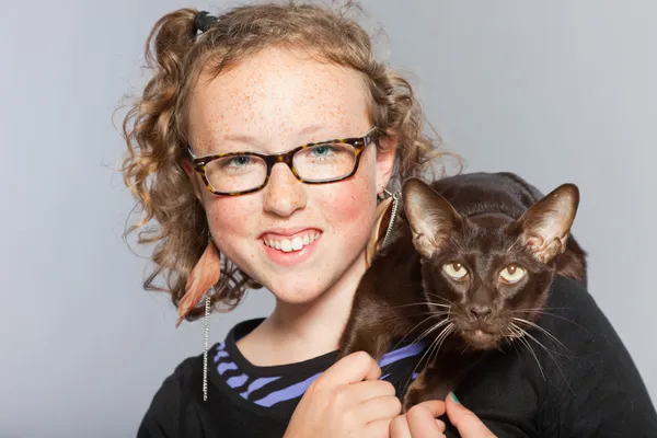 Happy teenage girl with glasses and blond curly hair hugging dark brown oriental shorthait cat.