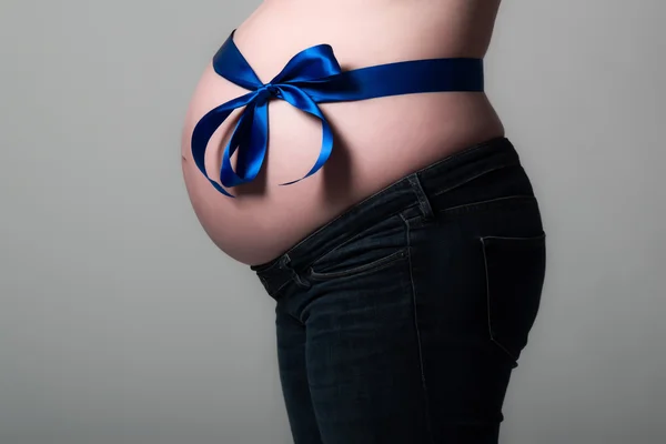 Pregnancy belly with blue ribbon isolated on grey. Wearing jeans