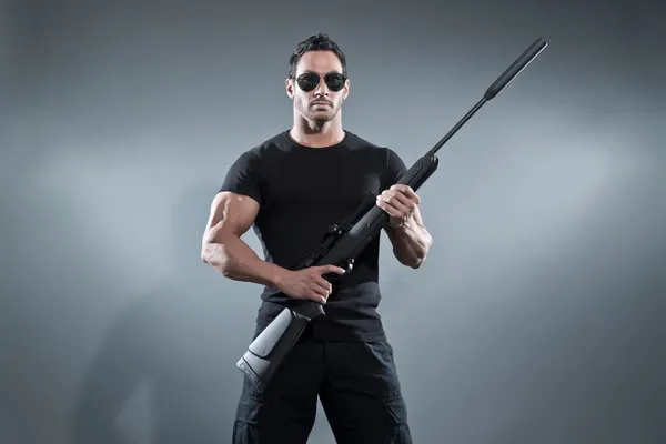 Action hero muscled man holding a rifle. Wearing black t-shirt w