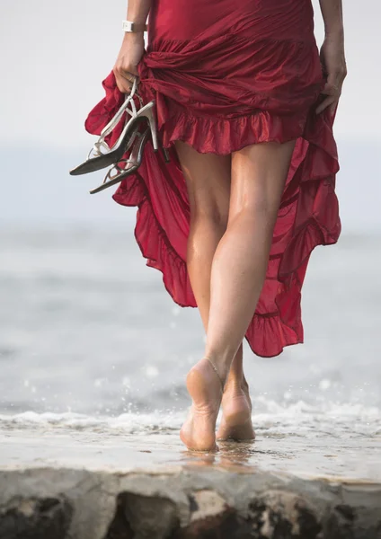 Sexy red dressed woman walks on wet stone next to sea