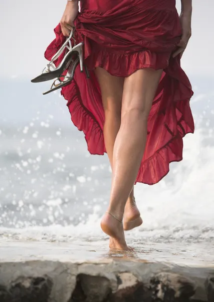 Sexy woman in red dress, steps on wet rock, next to sea