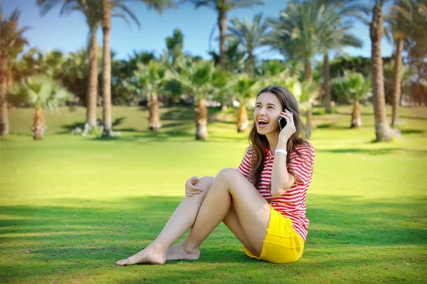Beautiful young woman sitting on a green lawn and talking on a cell phone