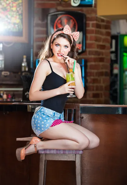 Attractive smiling pinup woman in denim shorts sitting on bar stool and drinking lemonade.