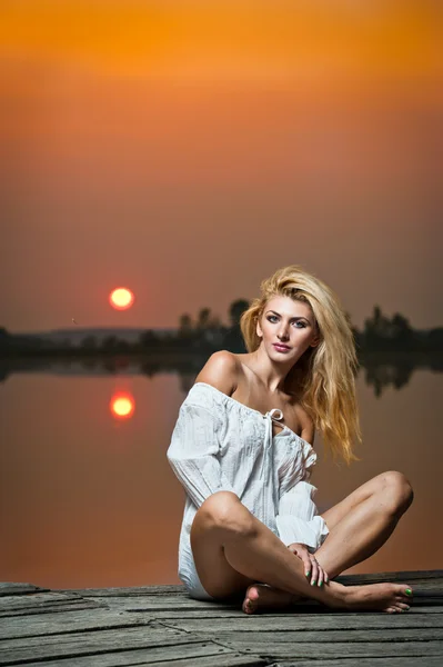 Beautiful girl with a white shirt on the pier at sunset.Sexy woman with long legs sitting on a pier .Color image of a beauty girl sitting on a pier, overlooking a lake