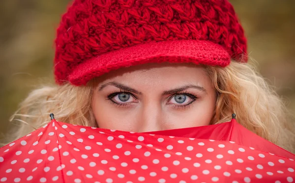 Beautiful blue eyes over colorful umbrella. Attractive blonde girl with red cap looking over red umbrella outdoor shoot. Attractive young woman in a autumn fashion shoot.