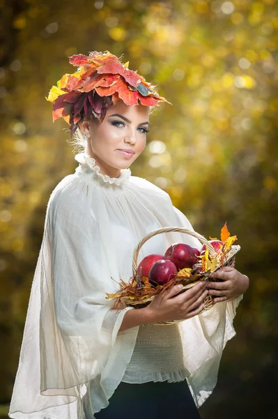 Beautiful creative makeup and hair style in outdoor shoot. Beauty Fashion Model Girl with Autumnal Make up and Hair. Fall. Beautiful fashionable girl with leaves in hair holding a basket with apples.
