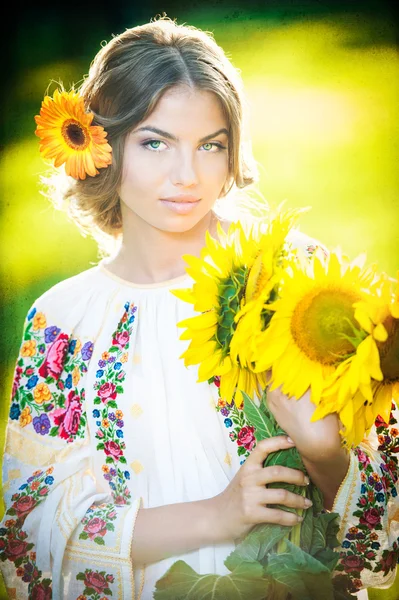 Young girl wearing Romanian traditional blouse holding sunflowers outdoor shot. Portrait of beautiful blonde girl with bright yellow flowers bouquet. Beautiful woman with yellow flower in hair posing