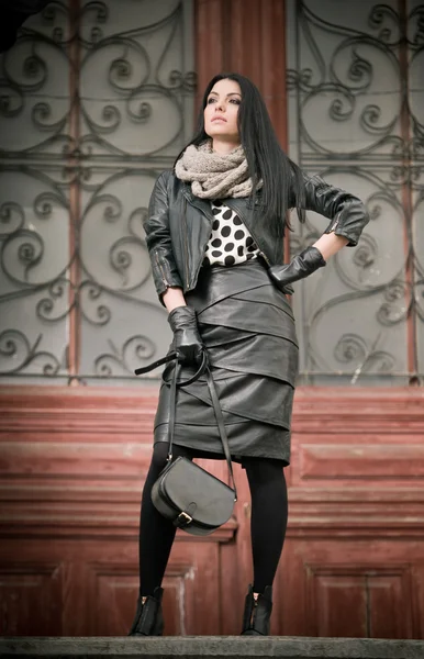 Attractive young woman in winter fashion shot with wrought iron decorated doors in background. Beautiful fashionable female - black leather outfit. Elegant long hair brunette posing in urban scenery.