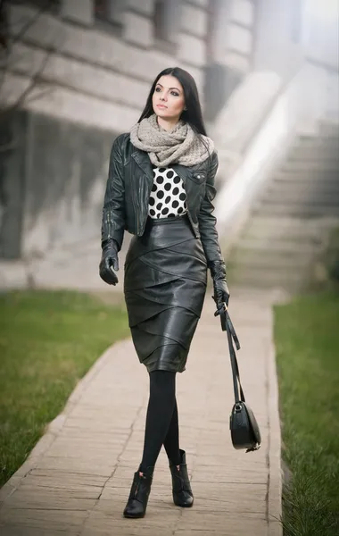 Attractive young woman in a winter fashion shot. Beautiful fashionable young girl in black leather waking on avenue. Elegant long hair brunette with handbag and scarf in urban scenery.