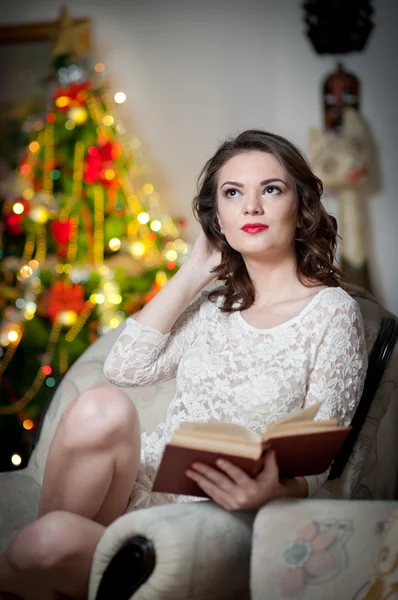 Beautiful sexy woman with Xmas tree in background reading a book sitting on chair. Portrait of a woman reading a book sitting comfortable with a blanket on legs. Attractive brunette female relaxing.