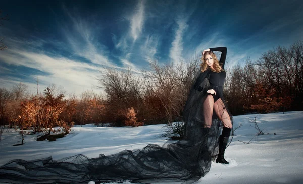 Lovely young lady posing dramatically with long black veil in winter scenery. Blonde woman with cloudy sky in background - outdoor shoot. Glamorous female in nature - gothic style