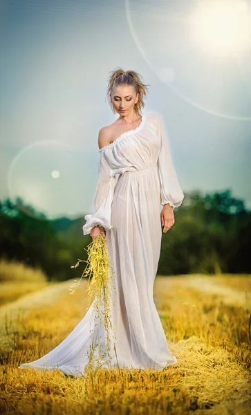 Young woman with long white dress standing on a wheat field. Portrait of girl outdoor. Romantic young woman posing on clean blue sky. Attractive woman in white dress in yellow wheat field at sunrise.