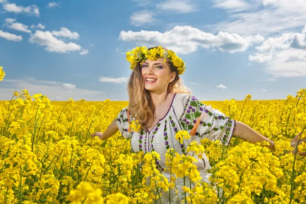 Young girl wearing Romanian traditional blouse posing in canola field with cloudy sky in background, outdoor shot. Portrait of beautiful blonde with flowers wreath smiling in rapeseed field