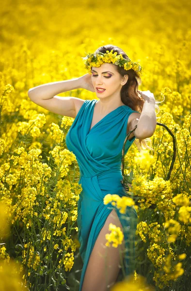 Fashion beautiful young woman in blue dress and yellow flowers wreath posing outdoor in canola field. Attractive long hair blonde girl with elegant dress smiling in rapeseed field in bright sunny day.