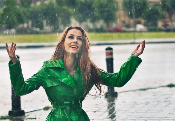 Beautiful woman in bright green coat posing in the rain. Happy redhead enjoying the rain drops in the park, outdoor shot. Beautiful red hair girl relaxing in a rainy summer day. Attractive female.
