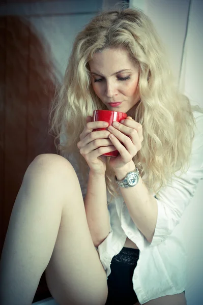 Portrait of a young, blond woman, holding a mug with both her hands, wearing a white shirt and black pants, with an expression of being sadness. Woman posing with a big red cup of tea in her hands.