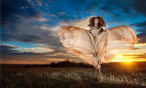 Fashionable beautiful young woman in nude colored long dress spinning around looking as a butterfly on cloudy dramatic sky in sunset. Attractive brunette with elegant luxurious dress, outdoors shot.