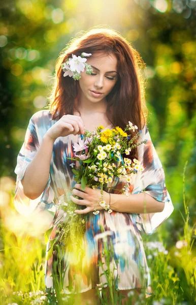 Young beautiful red hair woman holding a wild flowers bouquet in a sunny day. Portrait of attractive long hair female with flowers in hair, outdoor shot. Pretty girl enjoying the nature in summer
