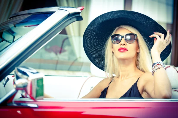 Outdoor summer portrait of stylish blonde vintage woman driving a convertible red retro car. Fashionable attractive fair hair female with black hat in red vehicle. Sunny bright colors, outdoors shot.