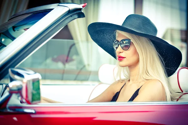 Outdoor summer portrait of stylish blonde vintage woman driving a convertible red retro car. Fashionable attractive fair hair female with black hat in red vehicle. Sunny bright colors, outdoors shot.