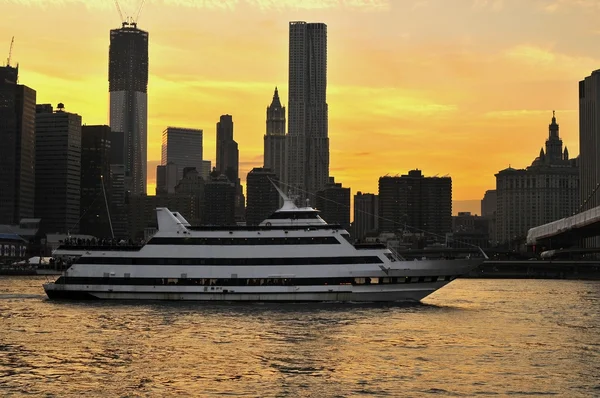 A river cruise boat on the East River heading under the Brooklyn Bridge in New York City at sunset time.